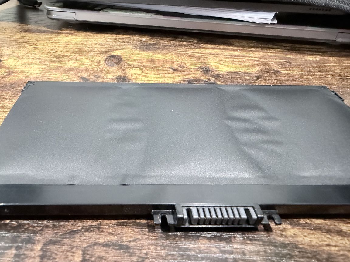 A swollen laptop battery with visible bulges resting on a workshop bench