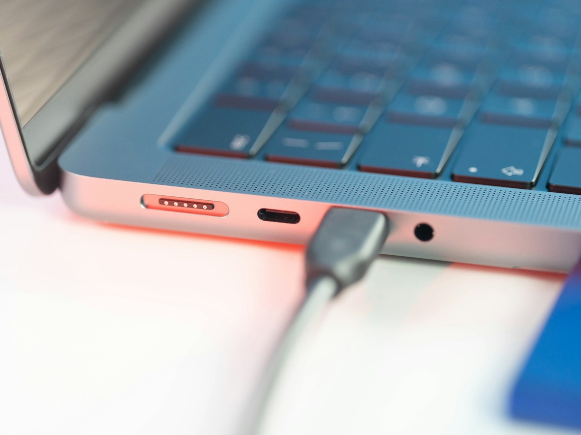 Close-up of a USB-C charging cable plugged into the side of a silver laptop, next to its magnetic charging port and headphone jack
