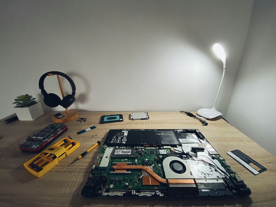 Laptop with its base cover removed on a wooden desk, showing the battery, cooling fan and motherboard, with a screwdriver set and removed parts beside it