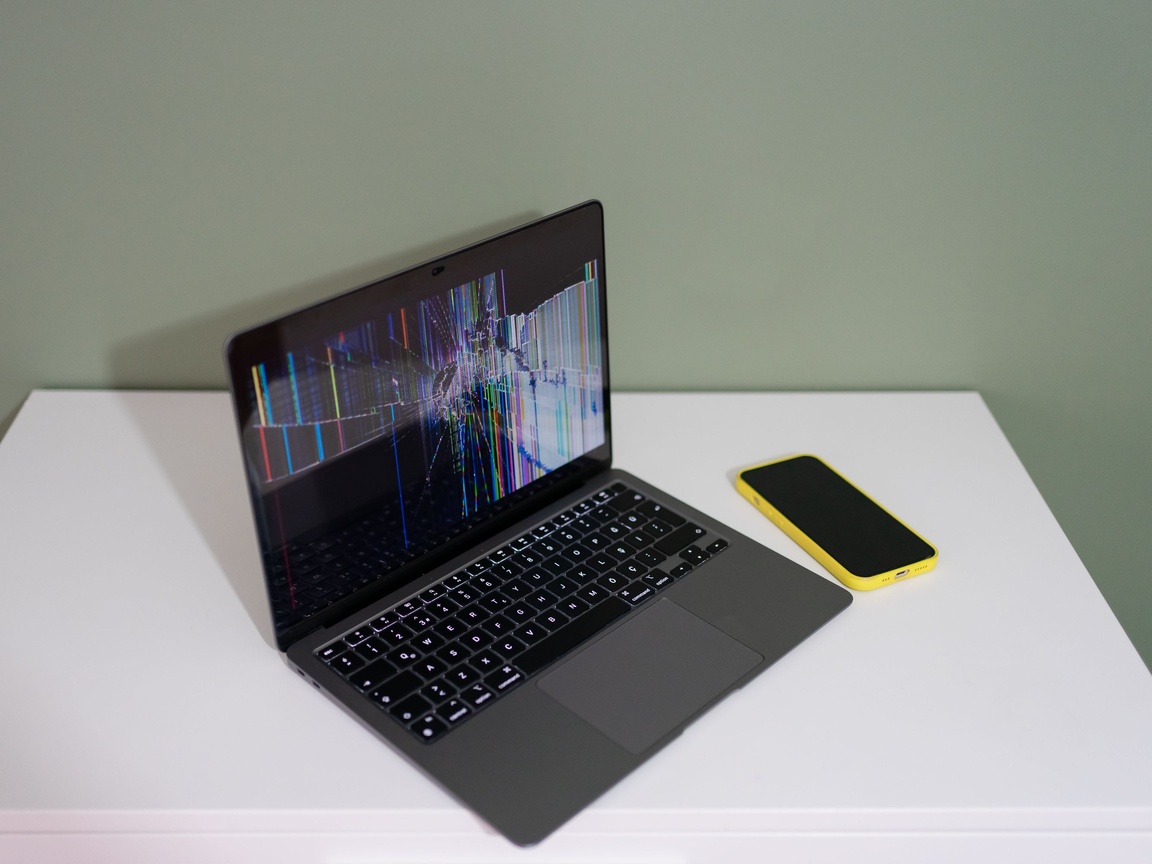 Laptop on a white table with a damaged screen showing coloured lines and cracks, next to a yellow smartphone